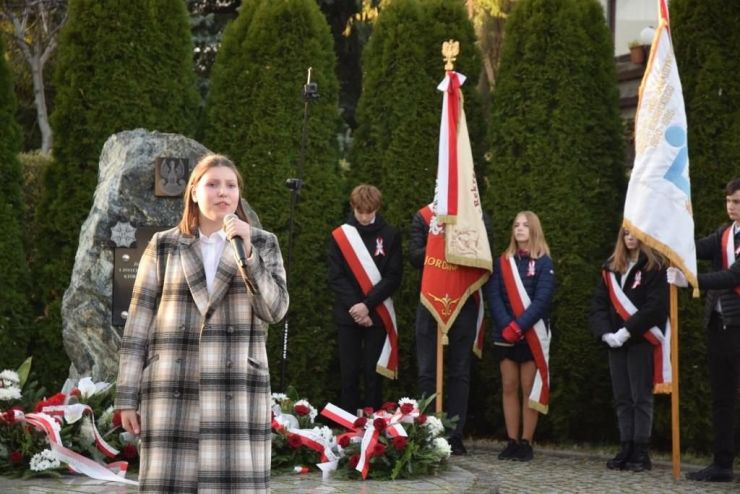 Nastolatka w beżowo czarnym płaszczu w kratkę trzyma mikrofon i śpiewa patriotyczną piosenkę. W tle obelisk i poczty sztandarowe szkół