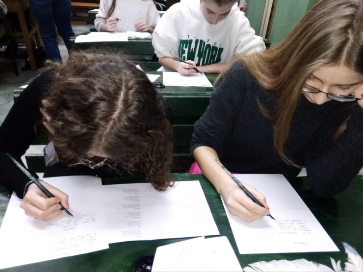 Uczniowie siedzą w zielonych ławkach szkolnych. Uczniowie trzymają w ręku pióro i piszą na kartce kaligrafią